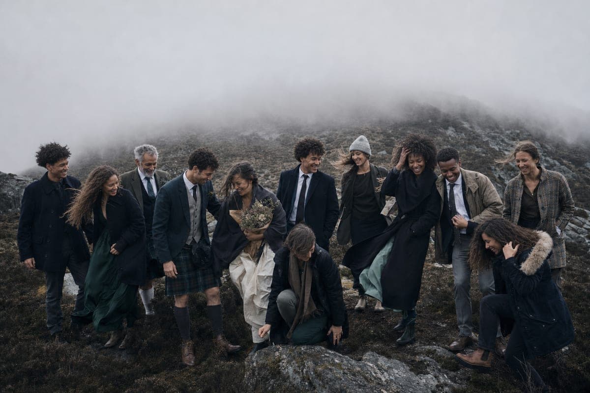 A wedding party crossing a heather hillside in low cloud, coats and hair blown sideways, one carrying wild flowers