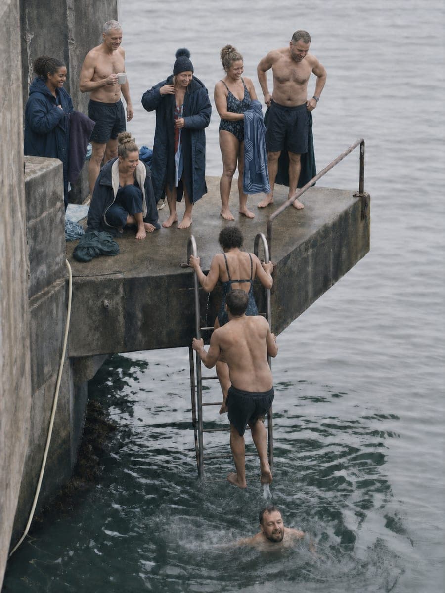 People on a concrete sea-bathing platform in winter, one climbing the ladder and one swimming below